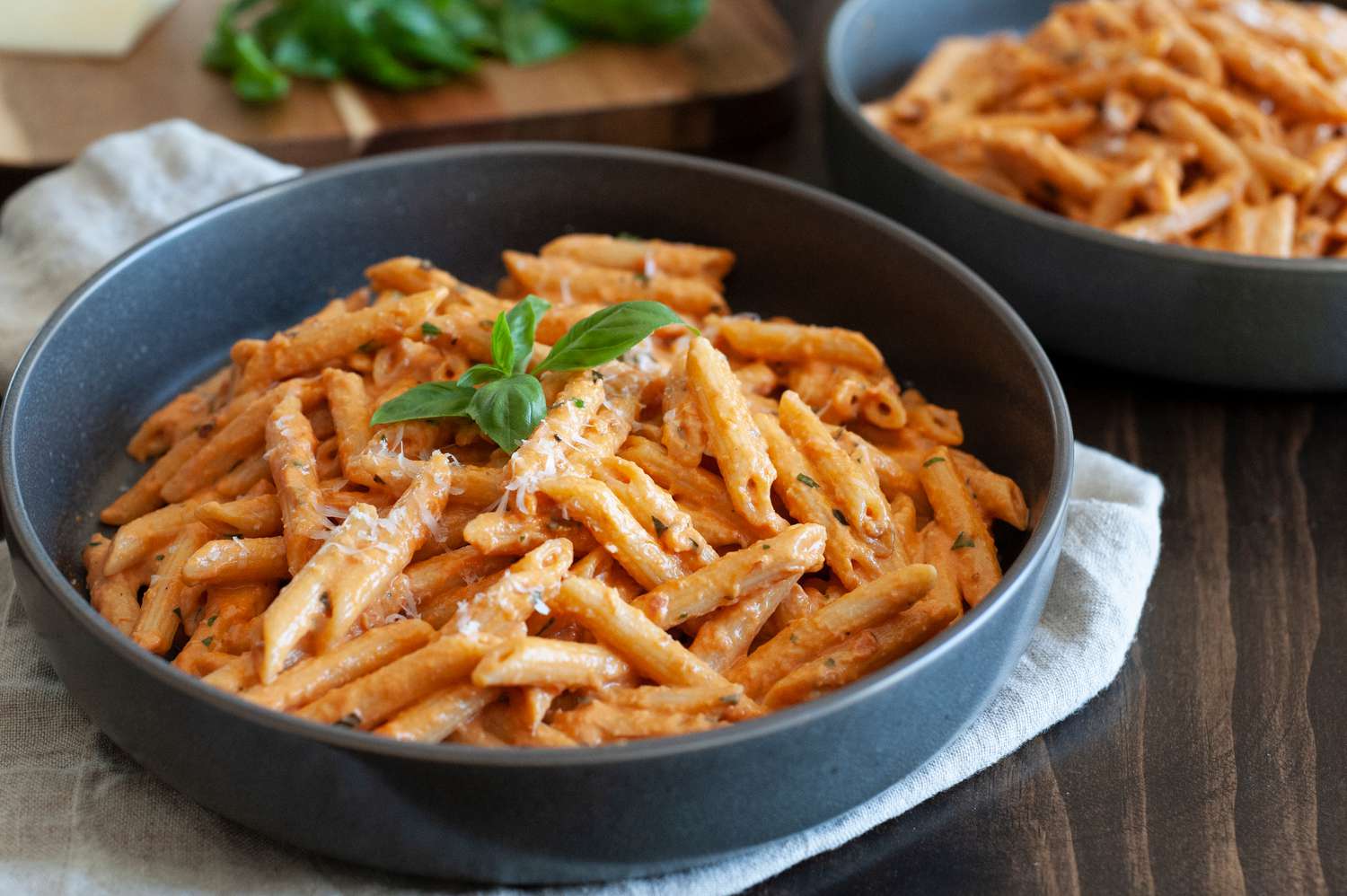 Penne alla Vodka in a Bowl Garnished with Basil over a Cloth Napkin. In Background, Another Bowl of Pasta and Cutting Board with Basil.