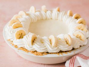 Whole banana cream pie with whipped cream swirls on top and banana slices set against a pink background.