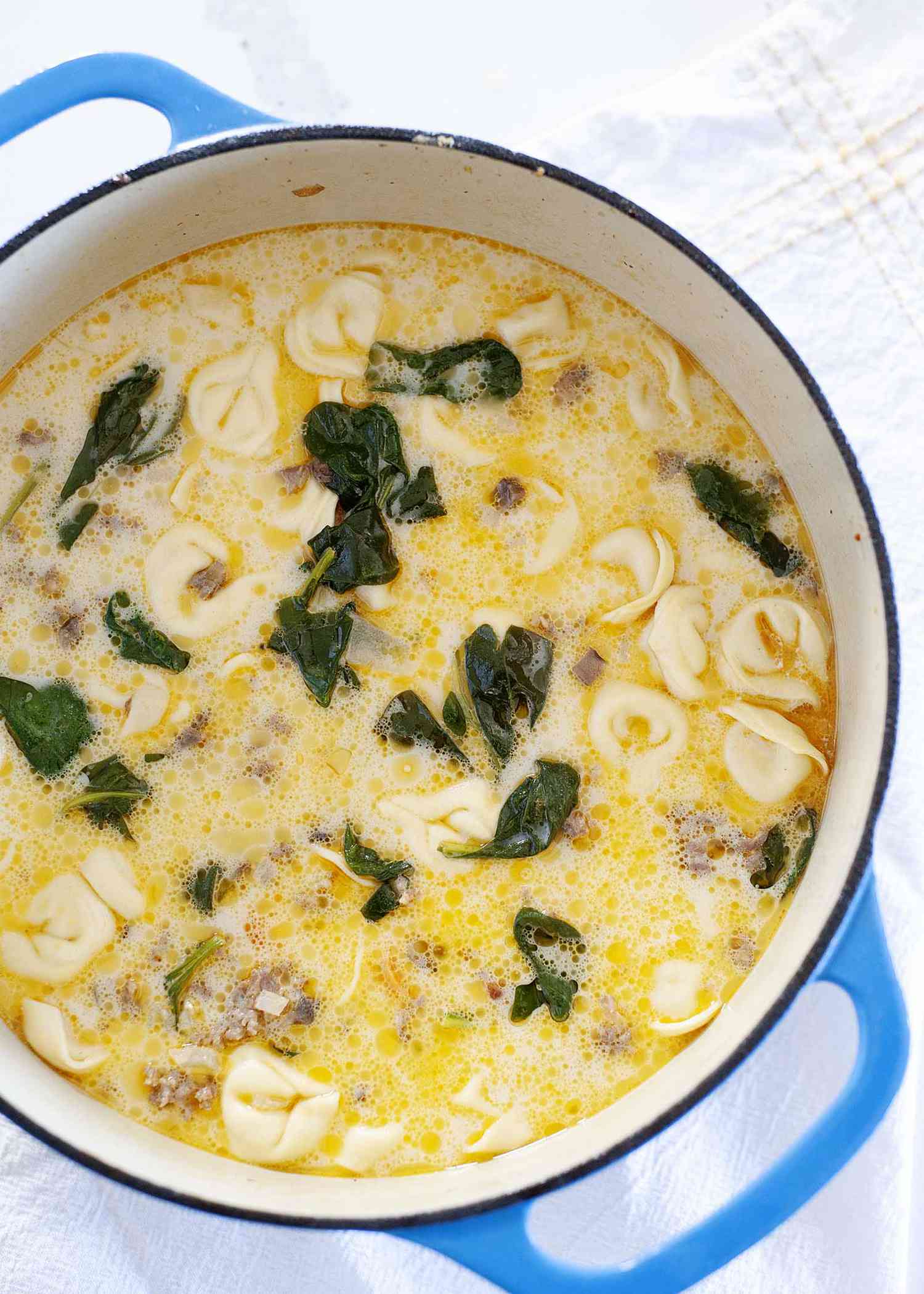 A blue dutch oven is filled with the best sausage tortellini soup and sits on a white tablecloth.