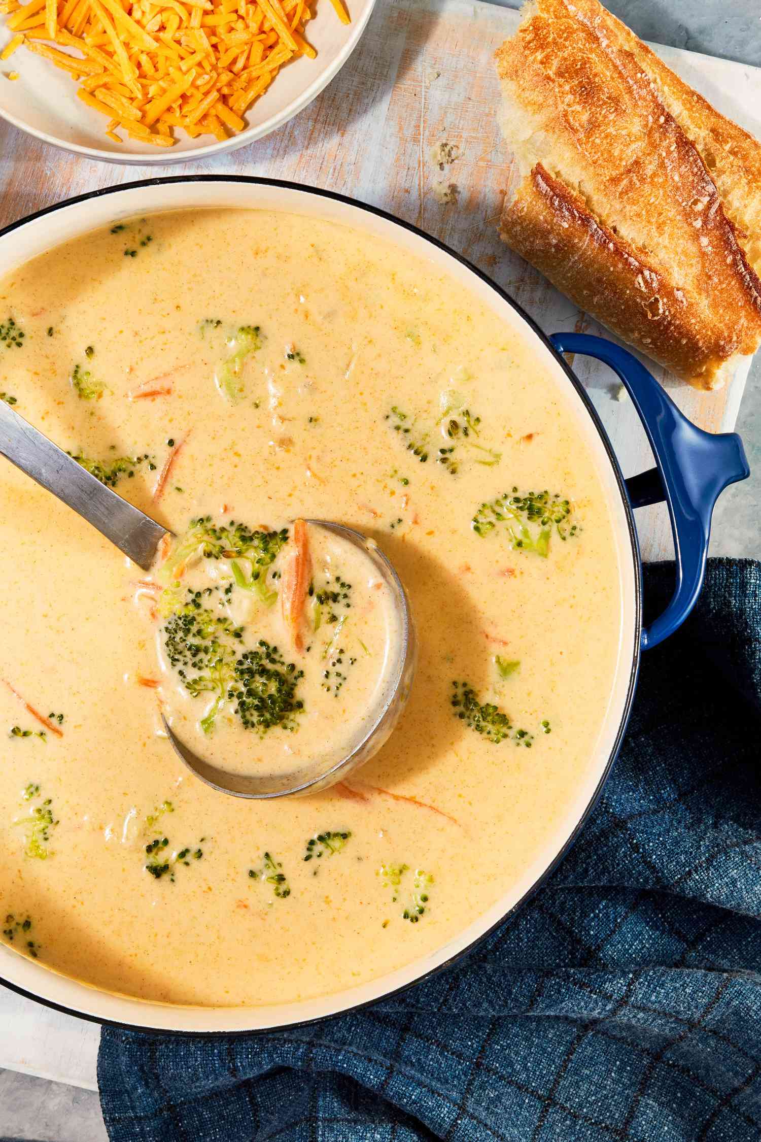 Pot of copycat Panera broccoli cheddar soup with a ladle raising some of it, and in the surroundings, a bowl of shredded cheese, a piece of baguette, and a blue kitchen towel