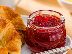 Closeup view of a small clear glass jar of strawberry jam on a plate with croissants and a knife all on a wooden tabletop