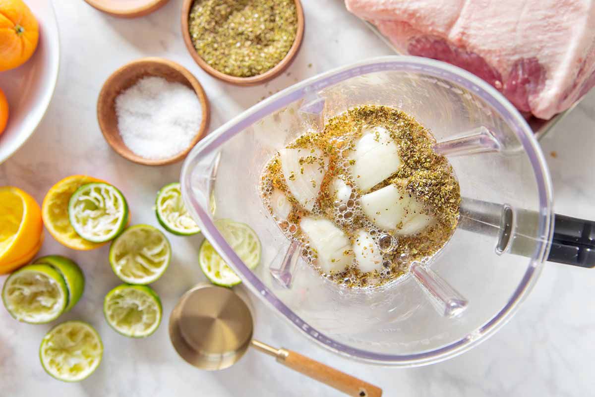 A clear blender pitcher has chopped onions, spices and liquid inside to make the best mojo marinated pork roast. Juiced limes, measuring cup, salt, spices and pork roast are set around the pitcher on a marble background.