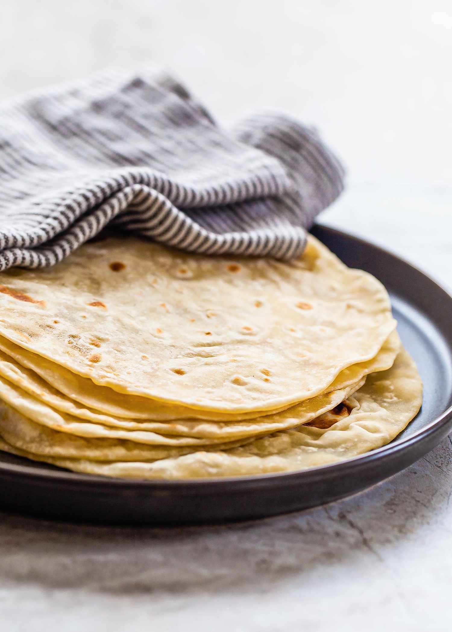 A close up of a stack of homemade flour tortillas on a platter covered by a striped towel.
