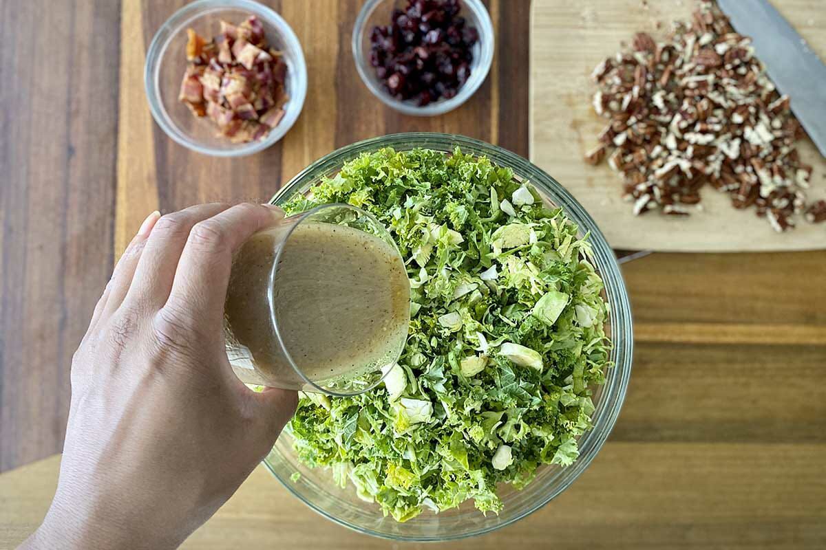 Pouring vinigrette over a bowl of shaved brussel sprouts and kale to make a Kale and Brussels Sprout Salad with Dried Cranberries and Maple Dijon Vinaigrette.