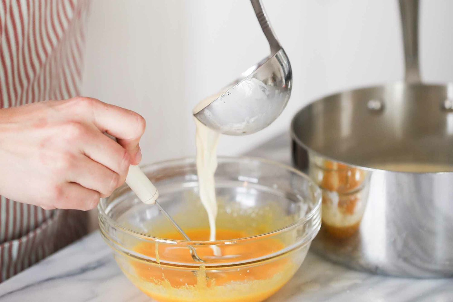 Tempering hot cream into eggs with a ladle next to a pot of cream