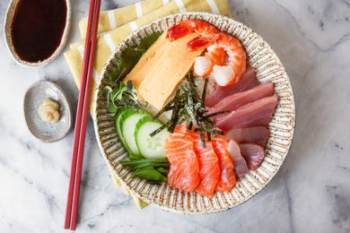 Chirashi Bowl with Salmon, Tuna, Shrimp, Tamagoyaki, and Sliced Cucumbers Next to a Small Saucer of Wasabi and Another Small Saucer of Soy Sauce