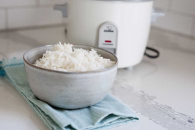 Horizontal image of a ceramic bowl filled with white rice on a white counter. A teal dish towel is under the bowl of rice and a white rice cooker is in partial view behind the bowl.