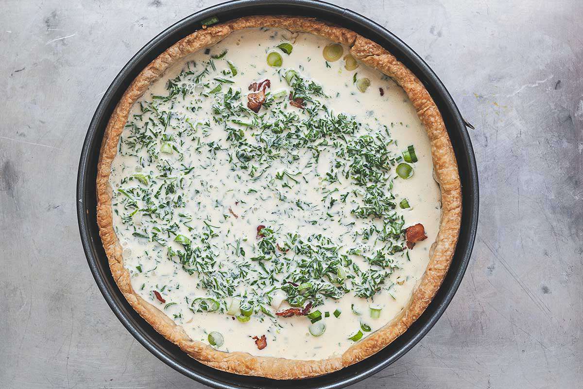 A homemade deep-dish bacon and cheddar quiche is on a countertop with an unbaked filling. The crust is golden brown and in a springform pan. Creamy filling with bacon, parsley and sliced scallionsns are visible in the crust.
