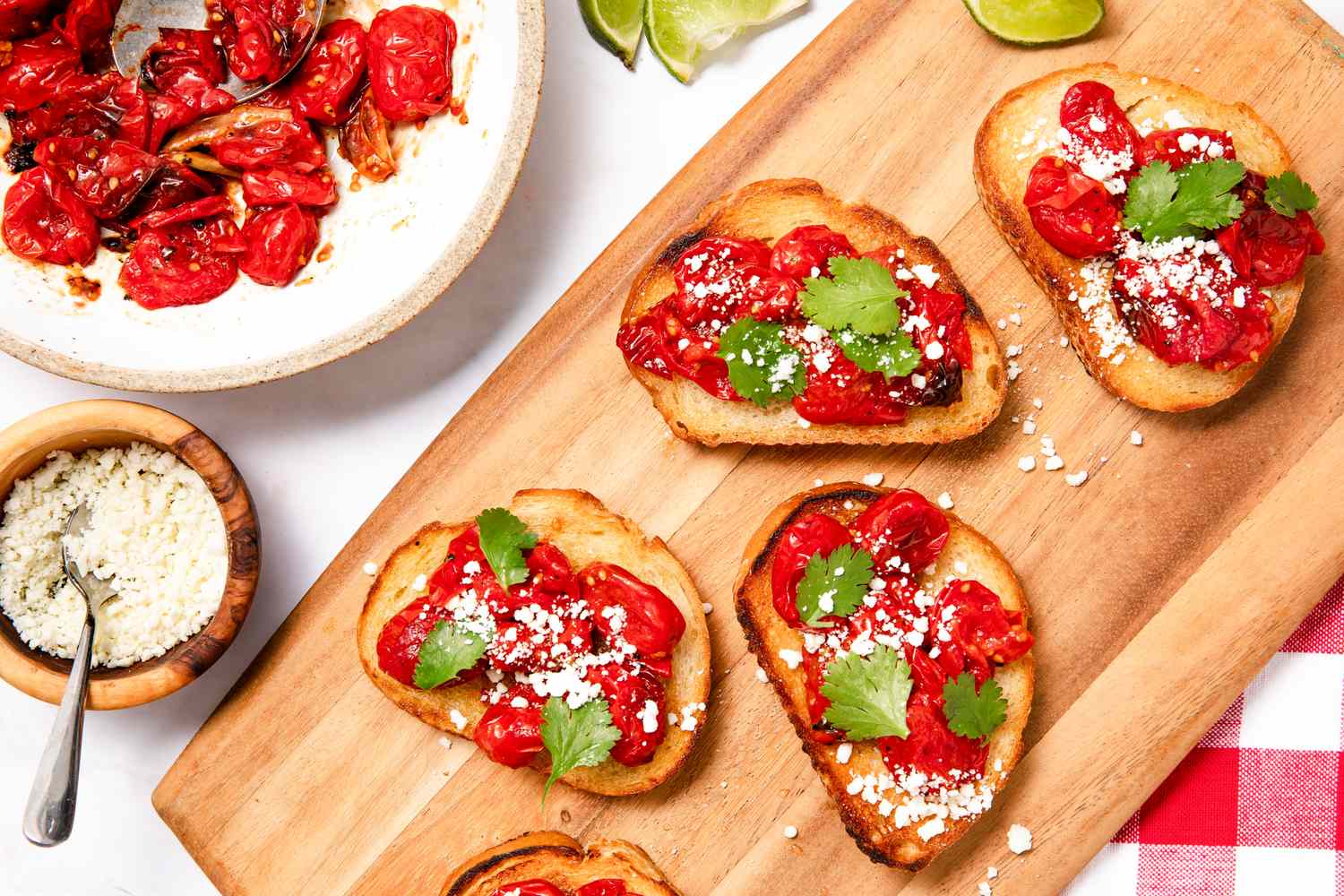 Blistered tomato toasts topped with fresh herbs and garnished with cheese, displayed on a wooden board alongside lime slices