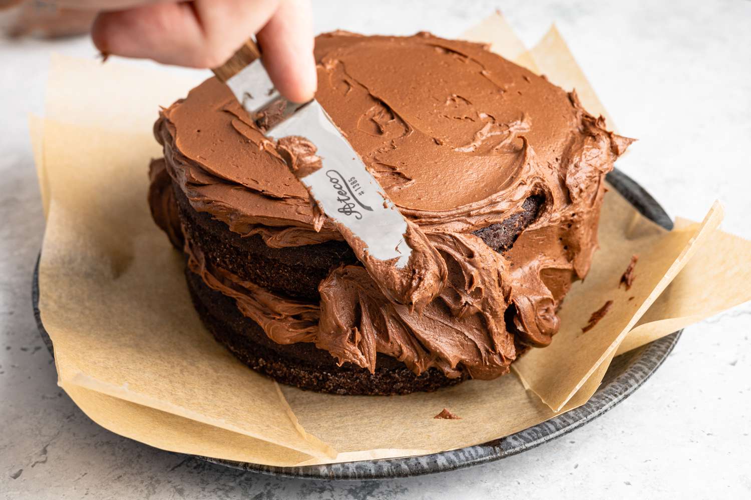 Offset Spatula used to Spread Chocolate Frosting Along the Sides of the Double-Layer Chocolate Cake on the Parchment Lined Plate