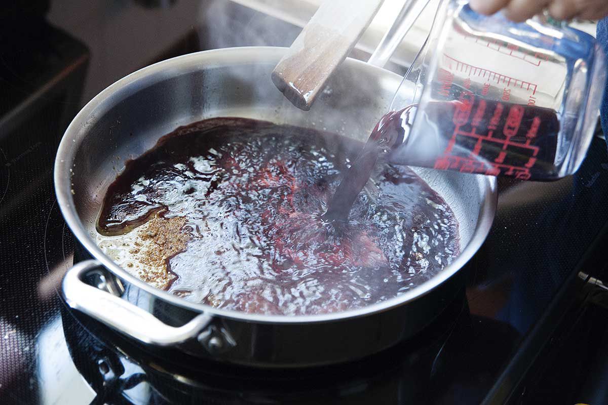 Pouring red wine into a a hot silver sauce pan to remove the fond searing a roast.