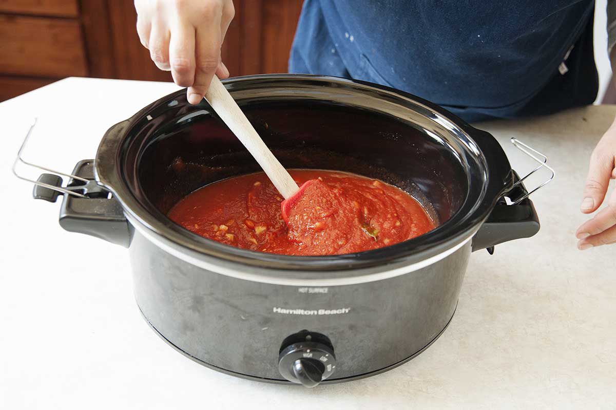 Tomato suace, herbs and onions being stirred together in a slow cooker for tomato sauce with slow cooker meatballs.