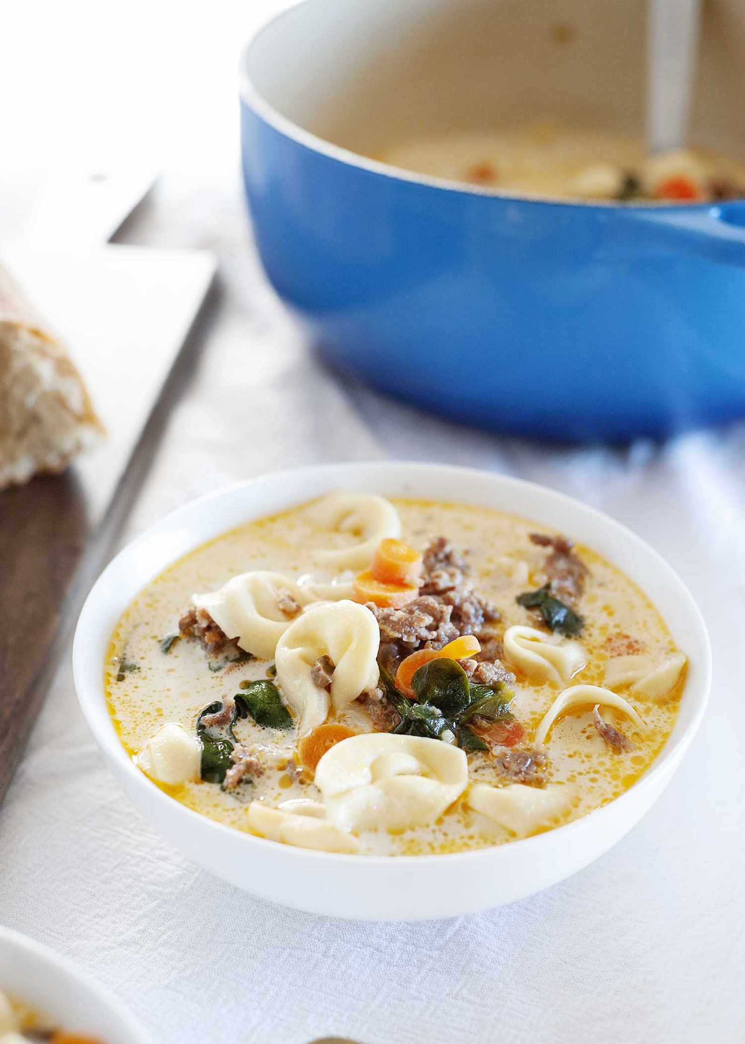Best homemade creamy tortellini soup in a white bowl on a table with a white tablecloth. A blue dutch oven with soup and spoon inside is behind the bowl.