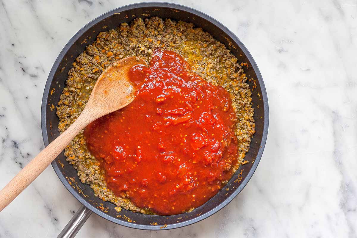 Meat and tomato sauce being stirred togehter with a wooden spoon in a skillet to make the perfect bolognese sauce.
