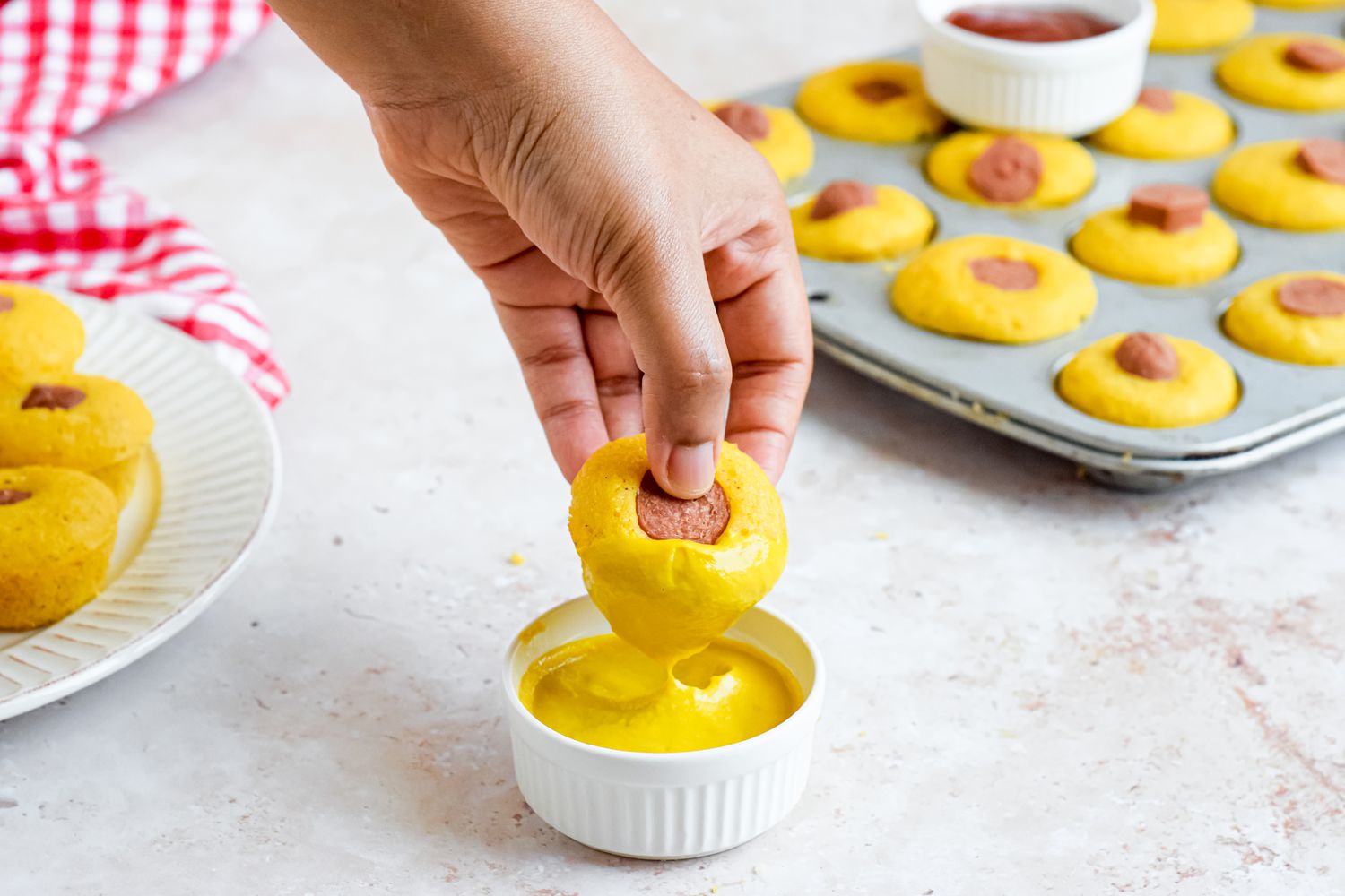 Hand Dipping a Corn Dog Muffin in a Bowl of Yellow Mustard, and in the Surroundings, a Plate With More Muffins, a Red and White Checkered Kitchen Towel, a Muffin Tin With More Muffin, and a Bowl of Ketchup Sitting on of the Muffin Tin