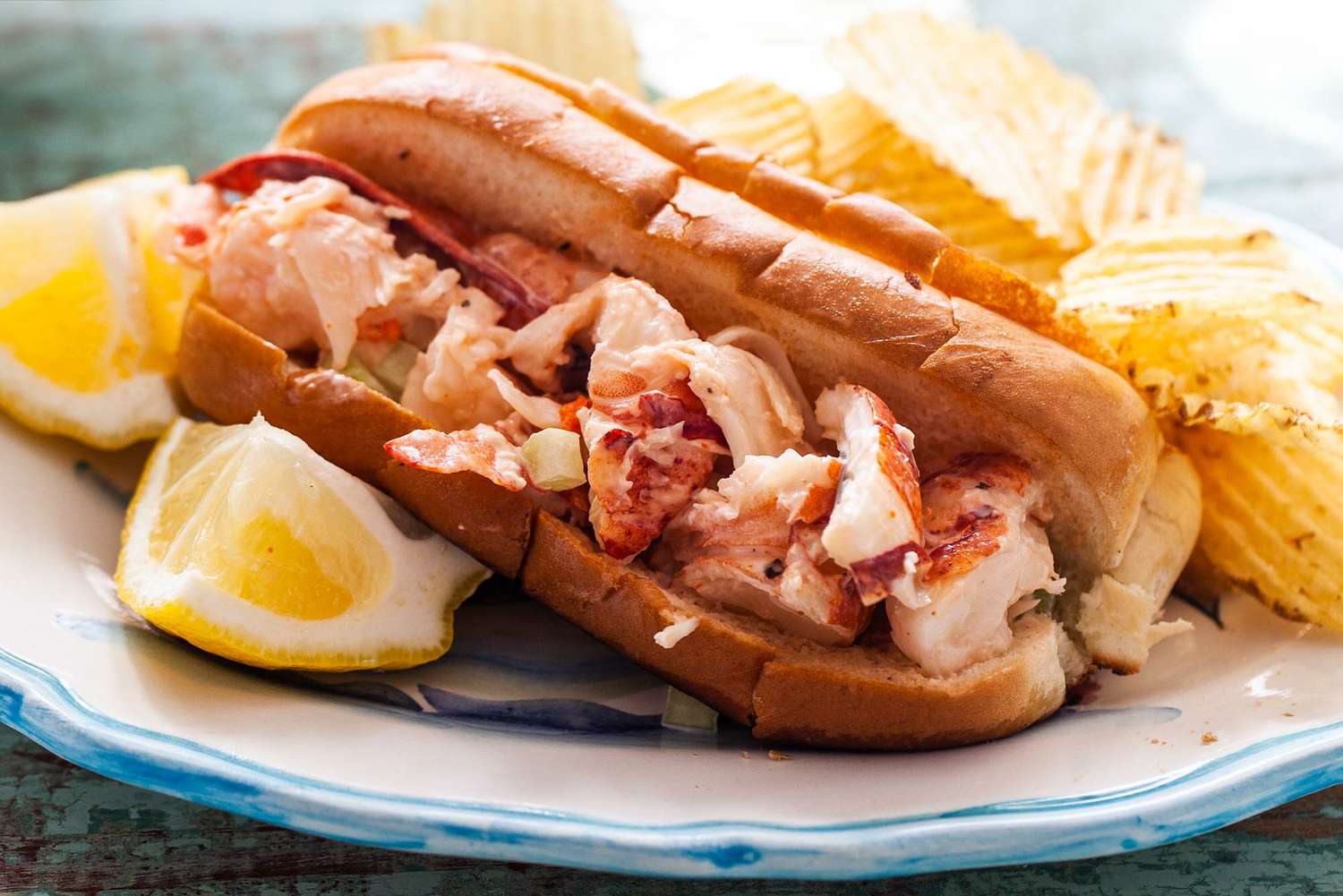 Lobster roll on a plate with potato chips and lemon wedges