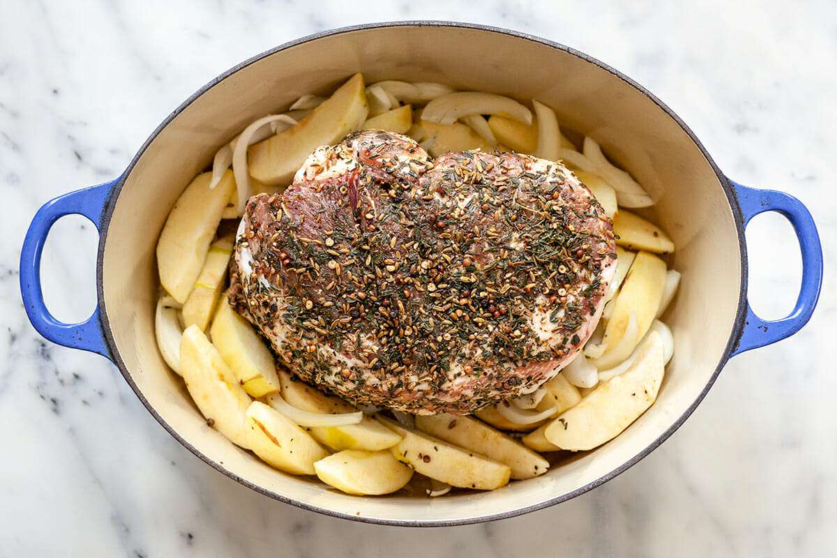 A dutch oven with a bone in pork shoulder roast set on top of apples and onions.