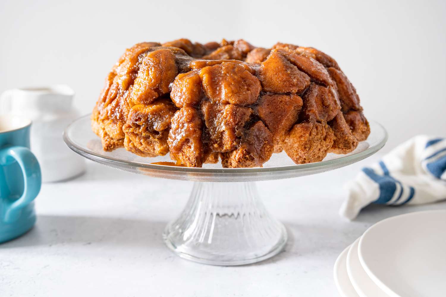 Monkey Bread on a Cake Stand, and on the Counter Surrounding It, a Stack of Plates, a Kitchen Towel, and a Coffee Mug