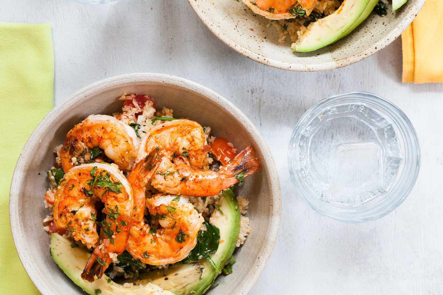 A horizontal poto of cauliflower rice and shrimp. A green napkin is to the left and a water glass to the right. Above the glass is a second bowl in partial view.