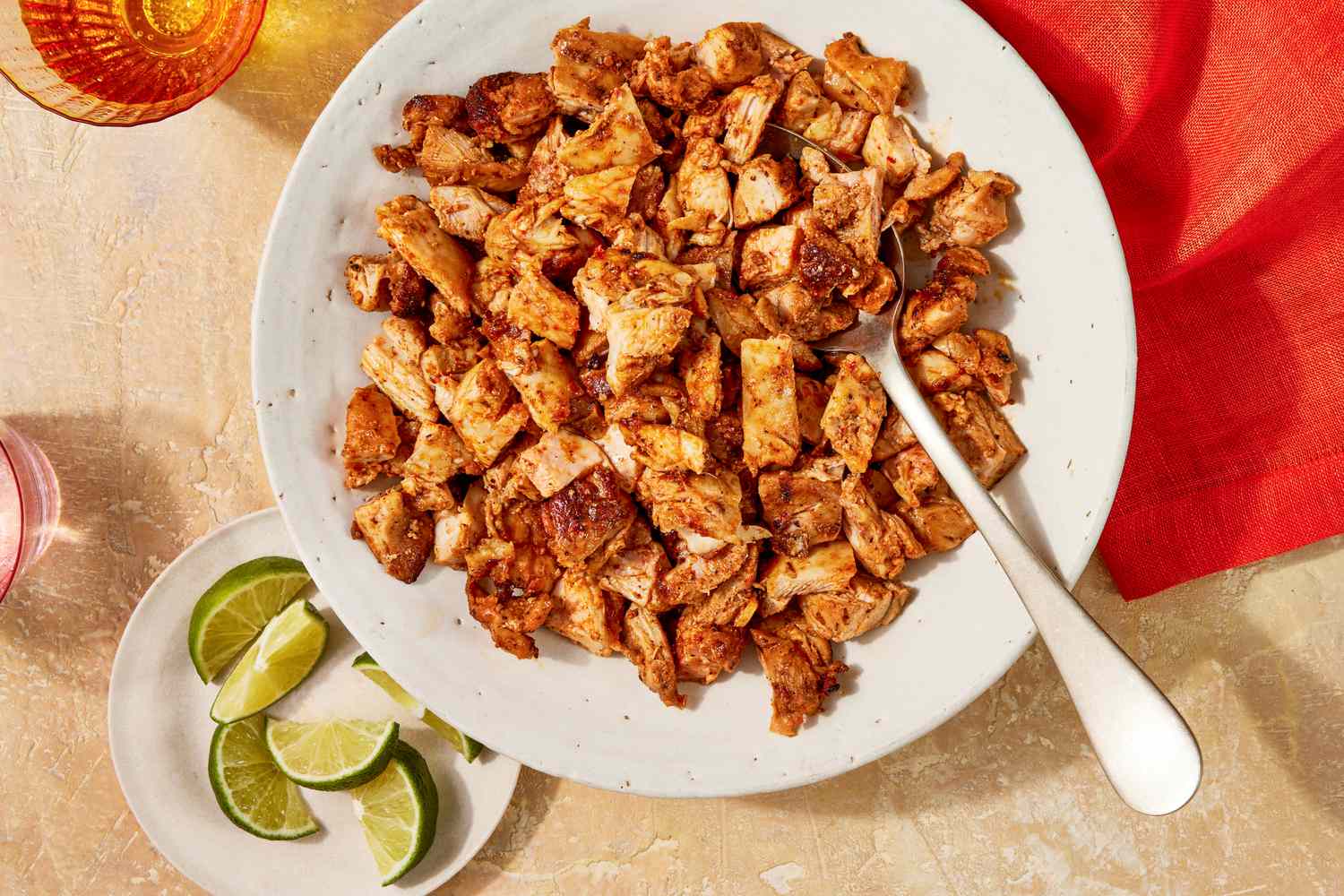 Bowl of copycat Chipotle chicken with a spoon, and in the surroundings, a bowl of lime quarters, glasses of water, and a scarlet table napkin