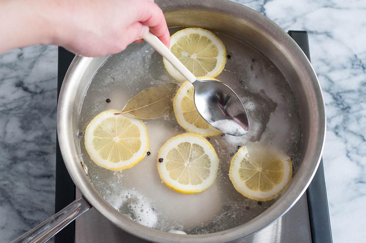 A hand holding a spoon and stirring a saucepan with poaching liquid and lemon slices