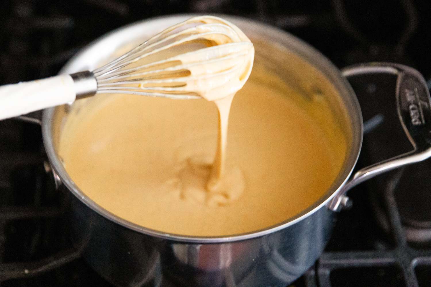 Angled view of a saucepan with a whisk after adding the final three cheeses simmering on a gas stovetop from Baked Mac and Cheese recipe