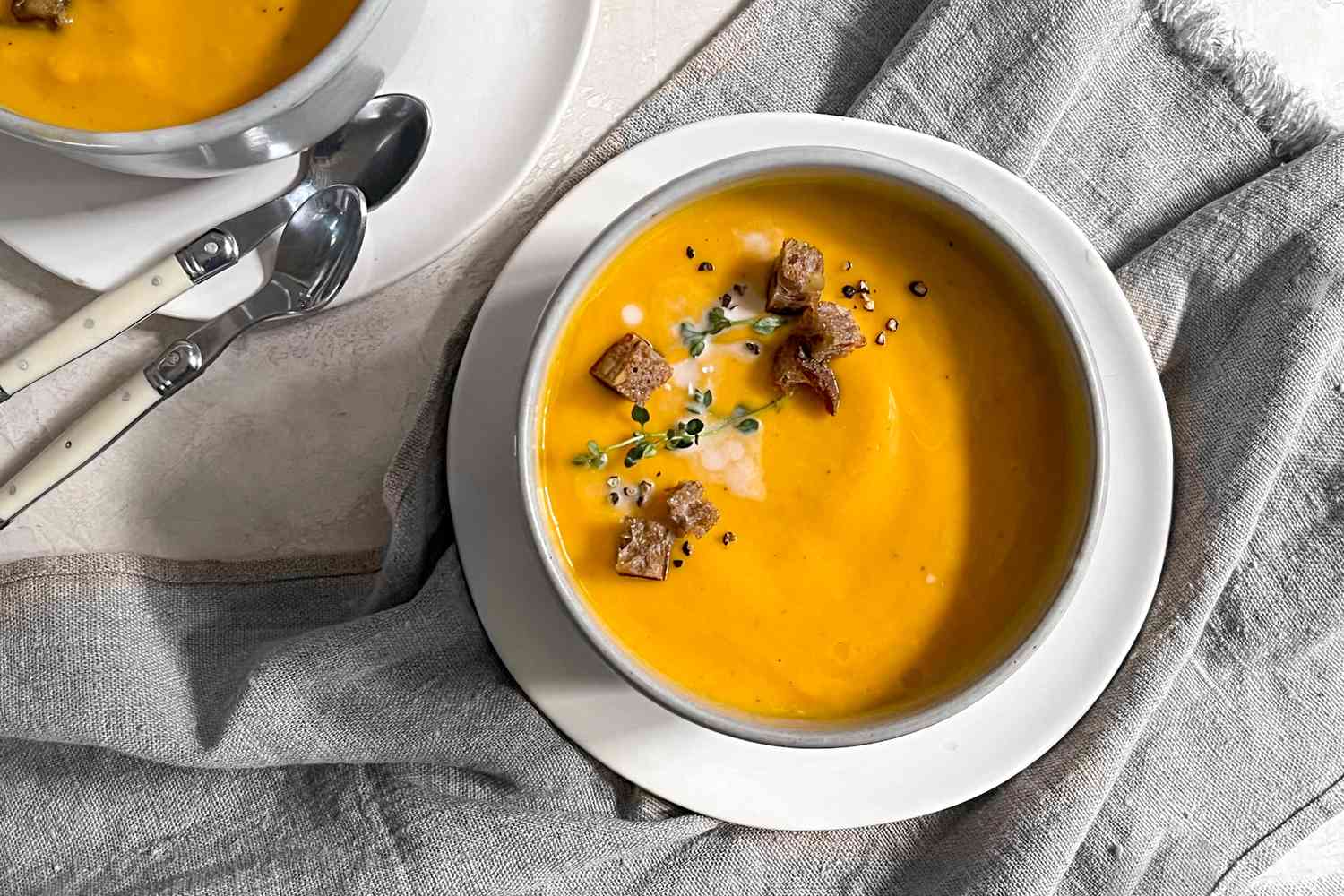 Bowl of Vegan Butternut Squash Soup Topped with Thyme, Coconut Milk, Croutons, and Cracked Pepper, Sitting on a Kitchen Linen and Next to Another Bowl of Soup and Spoons 