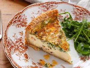 A slice of best deep dish quiche on a decorative china plate. A handful of arugula is to the right of the quiche. A fork is to the left of the plate and a table linen is above the plate.