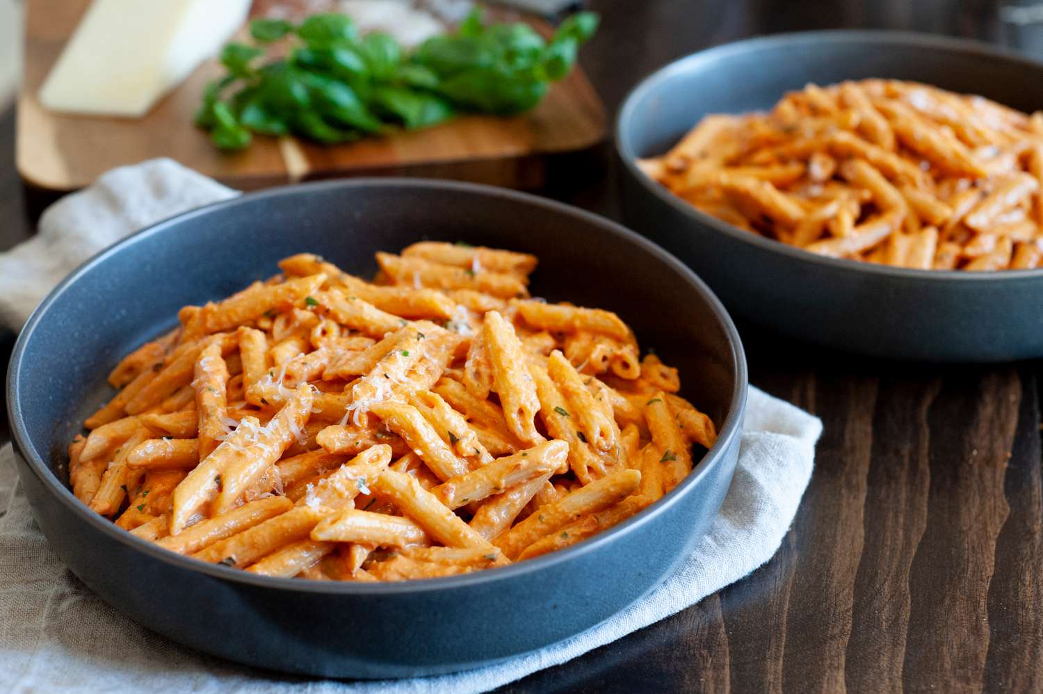 Penne alla Vodka in a Bowl over a Cloth Napkin, and in Background, Another Bowl of Pasta and Cutting Board with Basil and Cheese