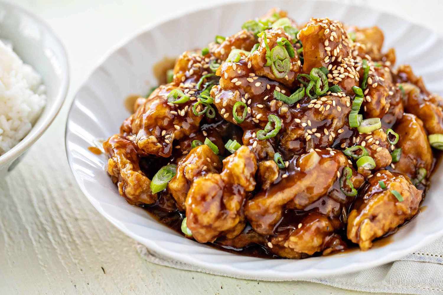 Side view of chinese take out orange chicken recipe in a scalloped white bowl with a partial view of a bowl of white rice to the left.