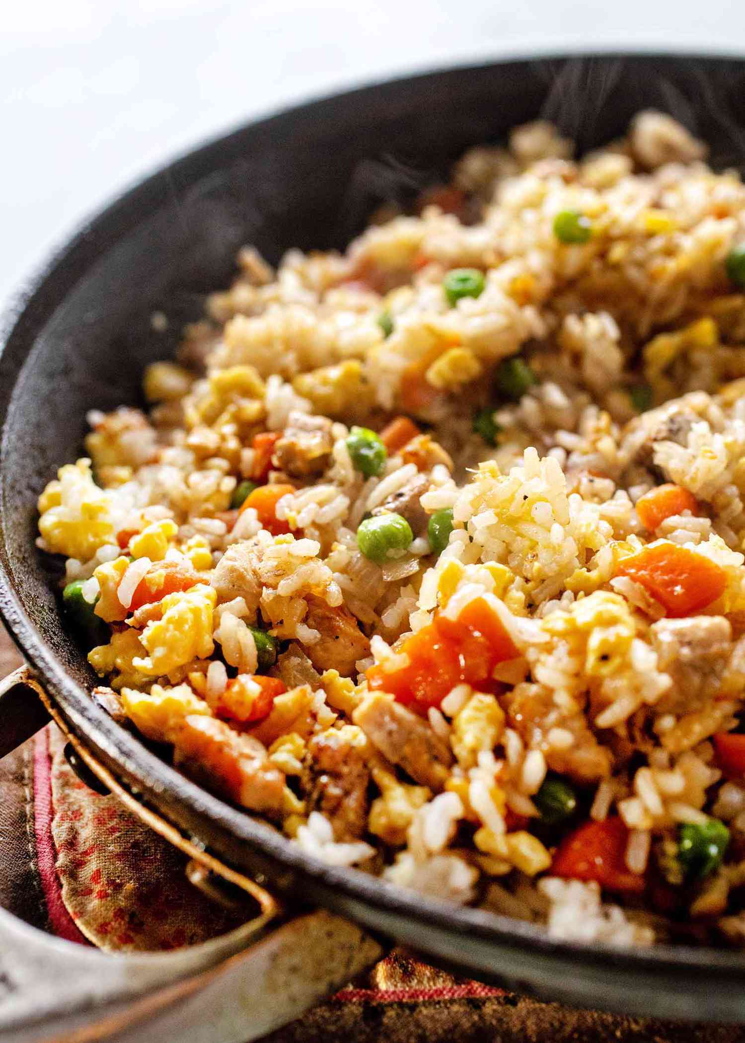Black skillet with pork fried rice recipe cooking inside. Carrots and peas are mixed in the rice.