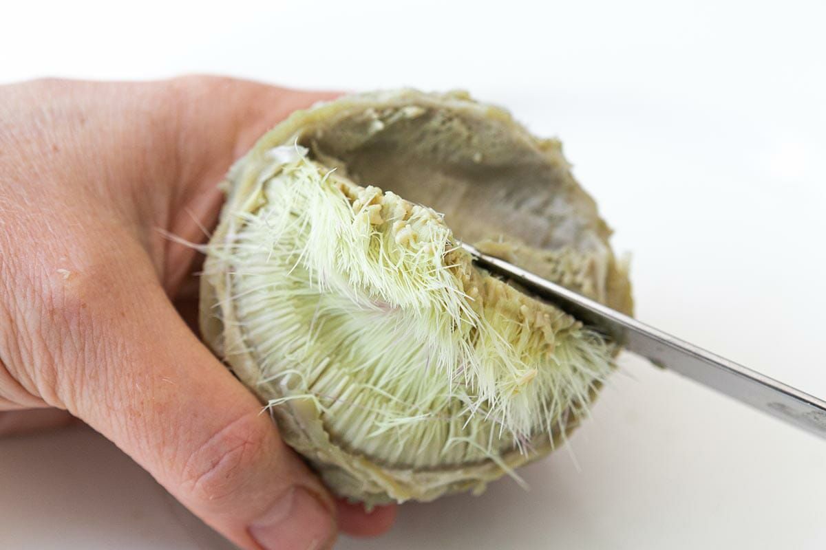 Hands holding a knife to scrape out and discard the choke from the very inside of a cooked artichoke heart