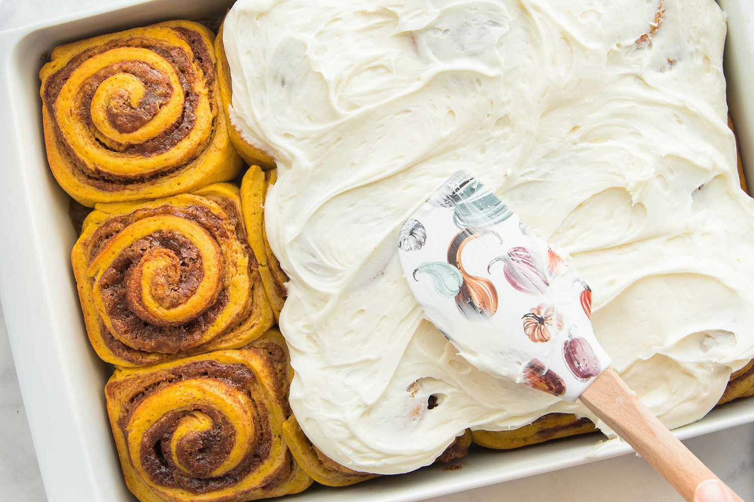 A pan of Homemade Pumpkin Cinnamon Rolls are being frosted.