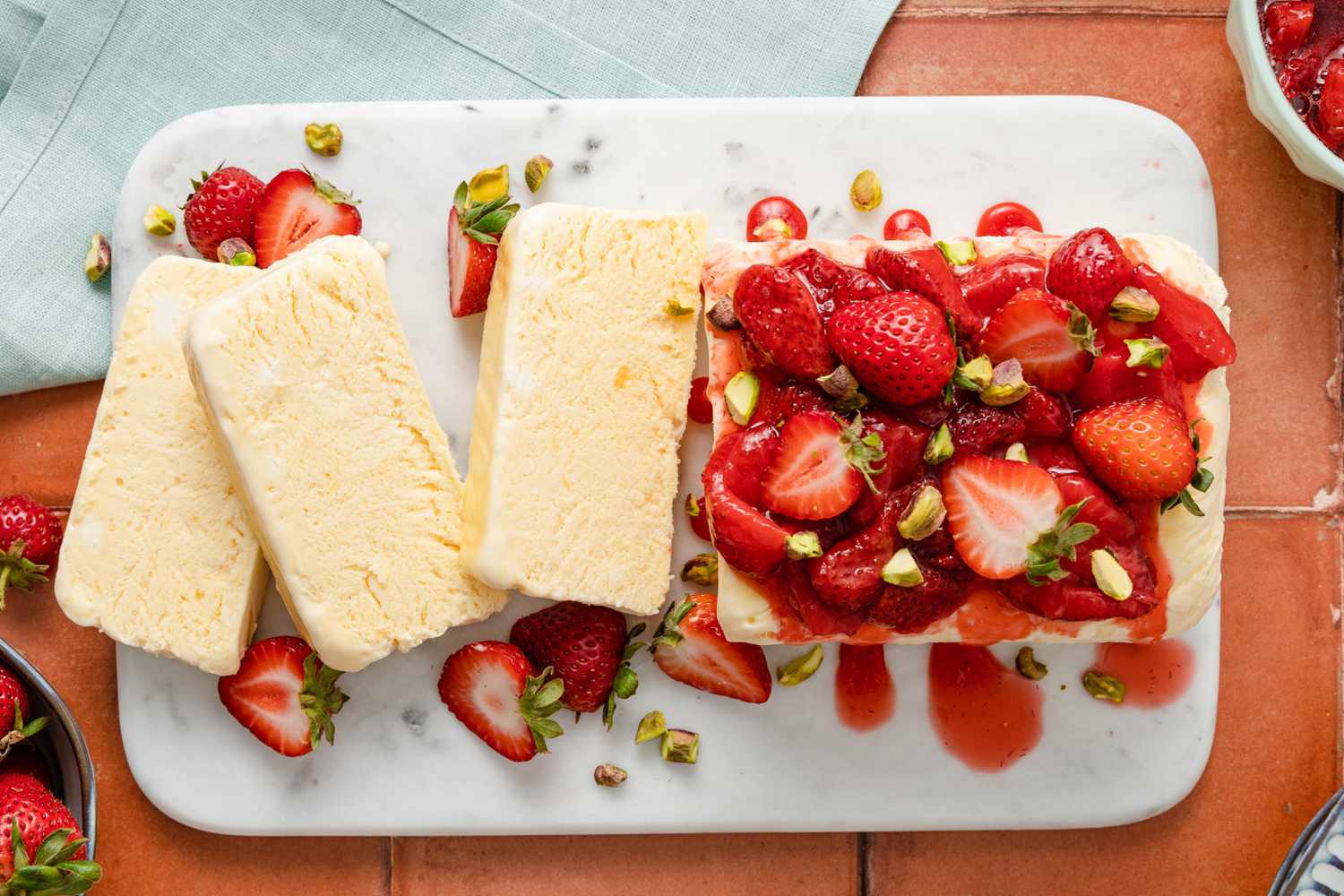 Overhead View: Strawberries and Cream Semifreddo Slices Cascading on a Platter Next the Rest of the Uncut Semifreddo on the Same Platter. Semifreddo Is Topped With Halved Fresh Strawberries and Strawberry Compote. In the Surroundings, a Bowl of Fresh Strawberries, a Stack of Plates, and a Blue Table Napkin.