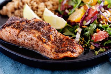 Side view of a fillet of broiled jerk salmon. The salmon has juices pooled at the bottom of it. A mixed green salad with citrus, two lime wedges and rice are visible on the plate behind the salmon. The plate is on a blue linen.