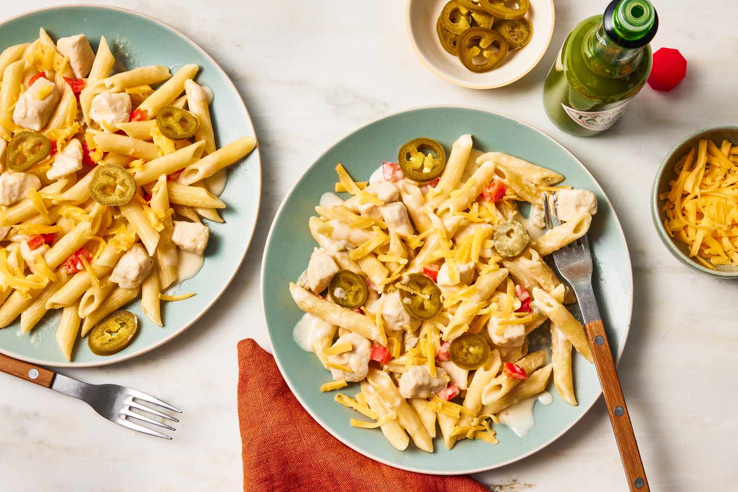 Overhead view of two light green plates of a chicken and penne pasta dish with cheese and jalapenos along with a fork and next to an orange napkin, bowl of jalapenos, bowl of cheese and a bottle of hot sauce on a beige background