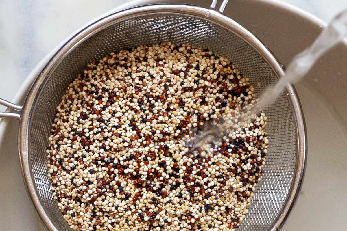 Uncooked quinoa in a colander with water being poured over top and draining in a bowl to show how to cook perfect quinoa.