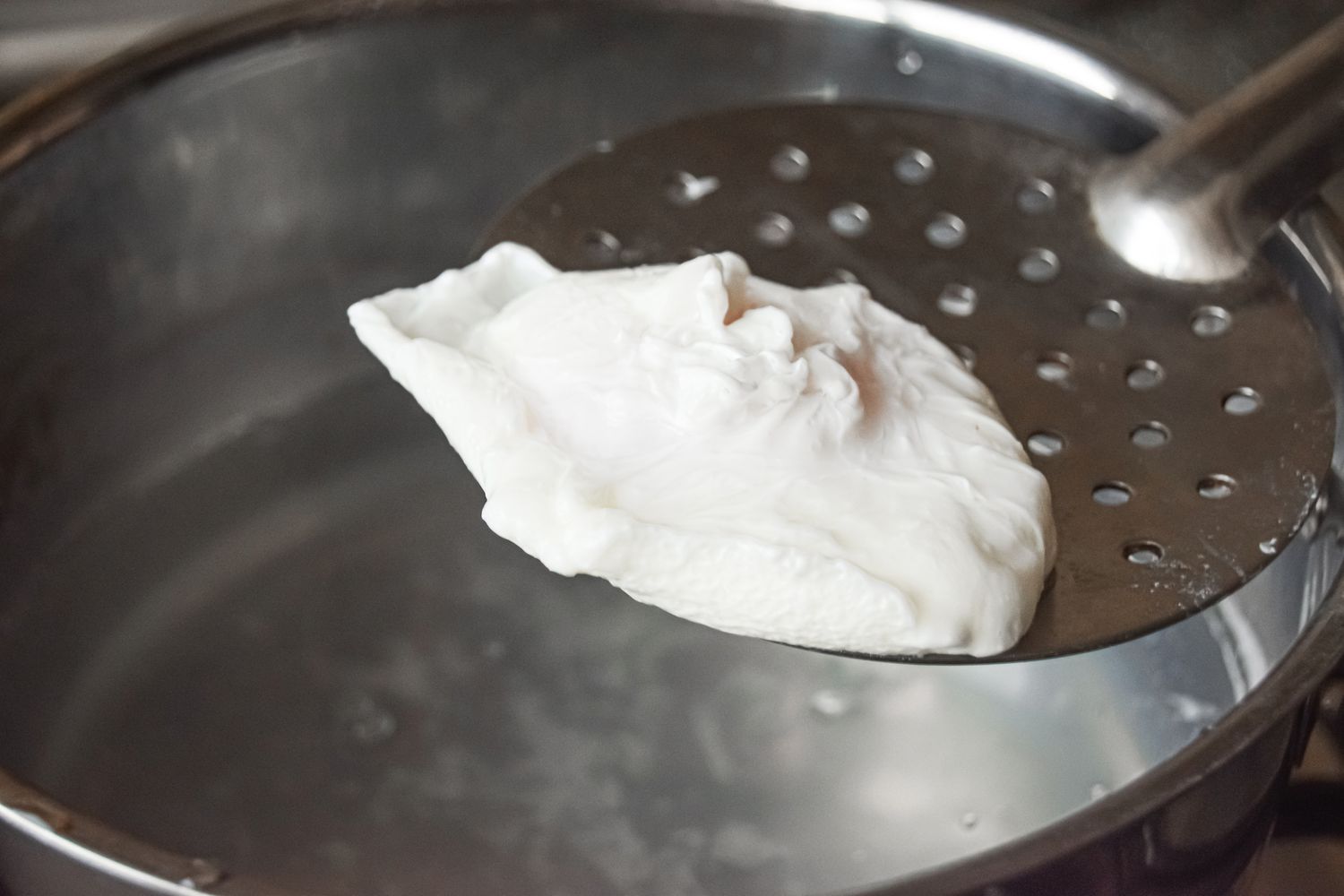 A slotted spoon lifting an egg out of a pot to show a poached egg