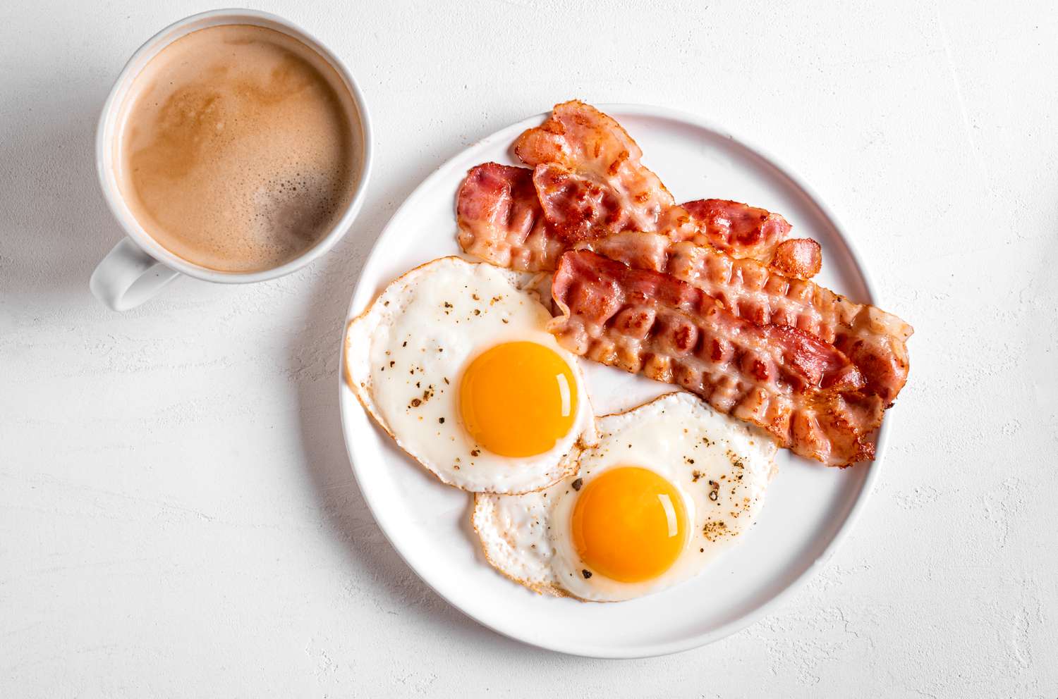 Two sunny side up eggs on a plate with bacon, next to a cup of coffee