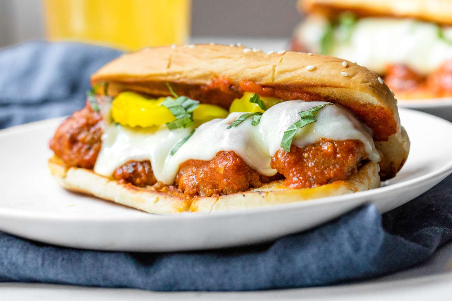 A Plate of Meatball Sub Topped with Banana Peppers and Basil, and in the Background, a Plate With Another Sub and a Glass of Beer