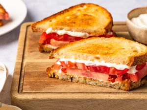tomato sandwich halves on a cutting board next to a bowl of mayo and a plate with more tomato sandwiches
