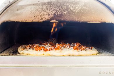 Roccbox pizza oven with a sausage pizza cooking inside. The flame is visible on the inside of the oven.
