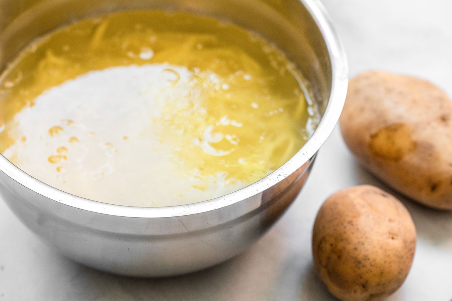 Grated Potatoes in a Bowl of Water Next to Some Uncut Potatoes for Air Fryer Hash Browns Recipe