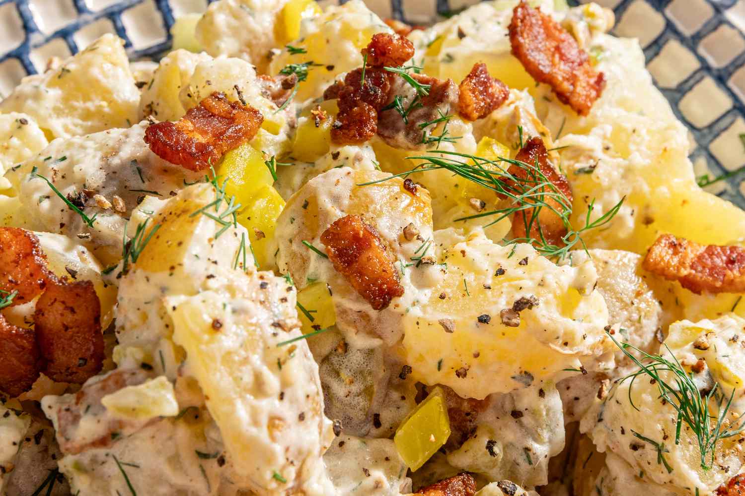 Potato salad with bacon pieces, herbs, and yellow pepper chunks served on a patterned background
