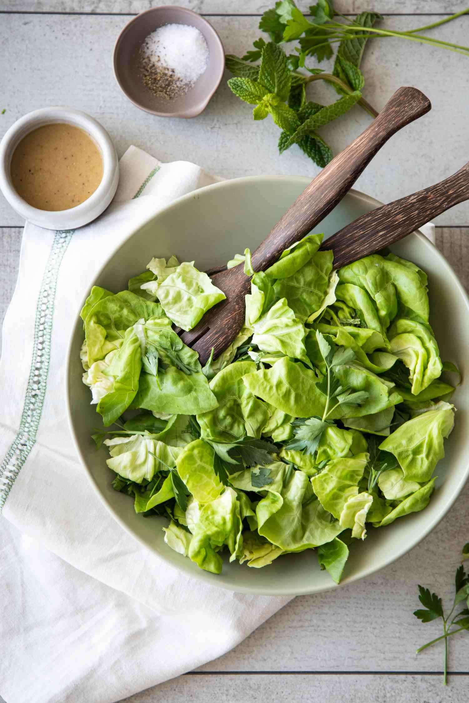 Large Bowl of Green Salad with Fresh Herbs with Serving Utensils Next Some Fresh Herbs, a Small Bowl of Red Wine Vinaigrette, and Another with Salt