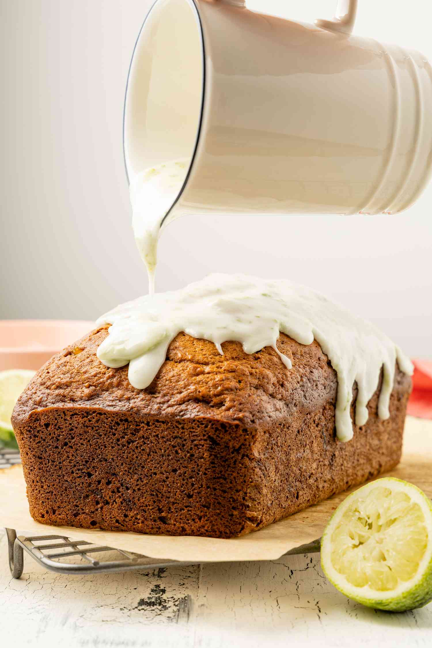 Pitcher of Lime Glaze Poured Onto a Loaf of Dark and Stormy Banana Bread on a Parchment Lined Cooling Rack, and on the Counter Next to It, a Halved Lime