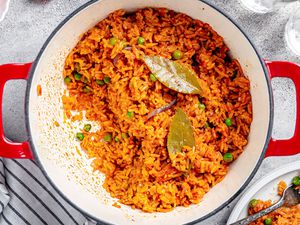 Jollof Rice in a Dutch Oven, and Next to It, a Plate With a Serving of Rice, a Small Saucer, and Glasses of Water