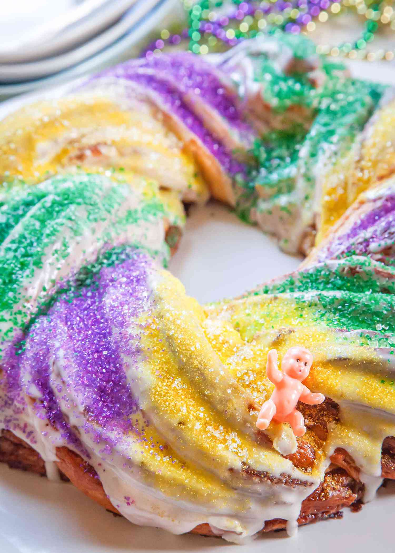 Side view of a homemade king cake on a white platter with a small plastic baby figurine in the bottom right hand corner.