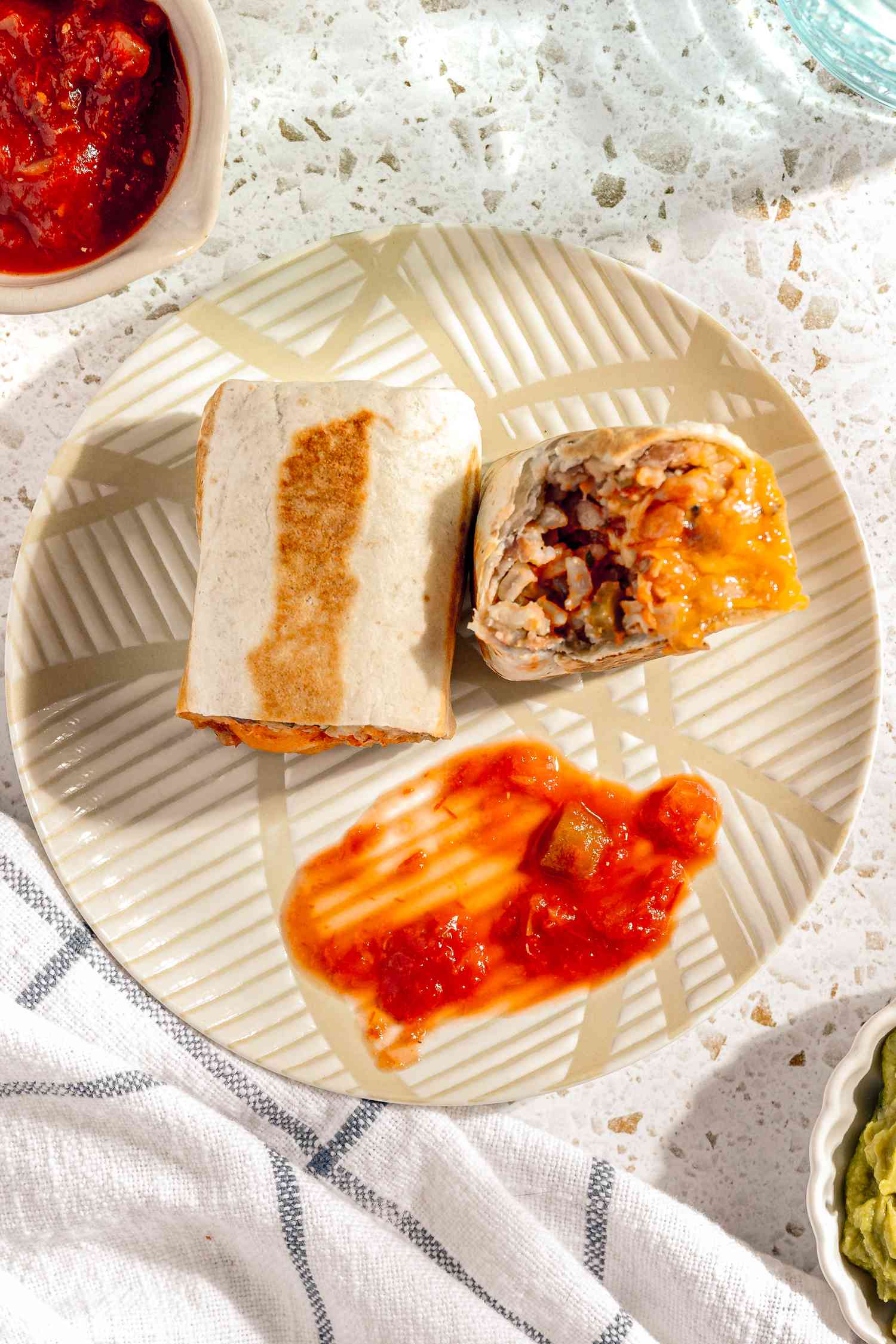 Easy Rice and Beans Freezer Burrito Cut in Half (One Half's Cross-Section Facing Camera) on a Plate and Served With Salsa, and in the Surroundings, a Small Bowl of Salsa, a Small Bowl of Guacamole, a Glass of Water, and a Kitchen Towel