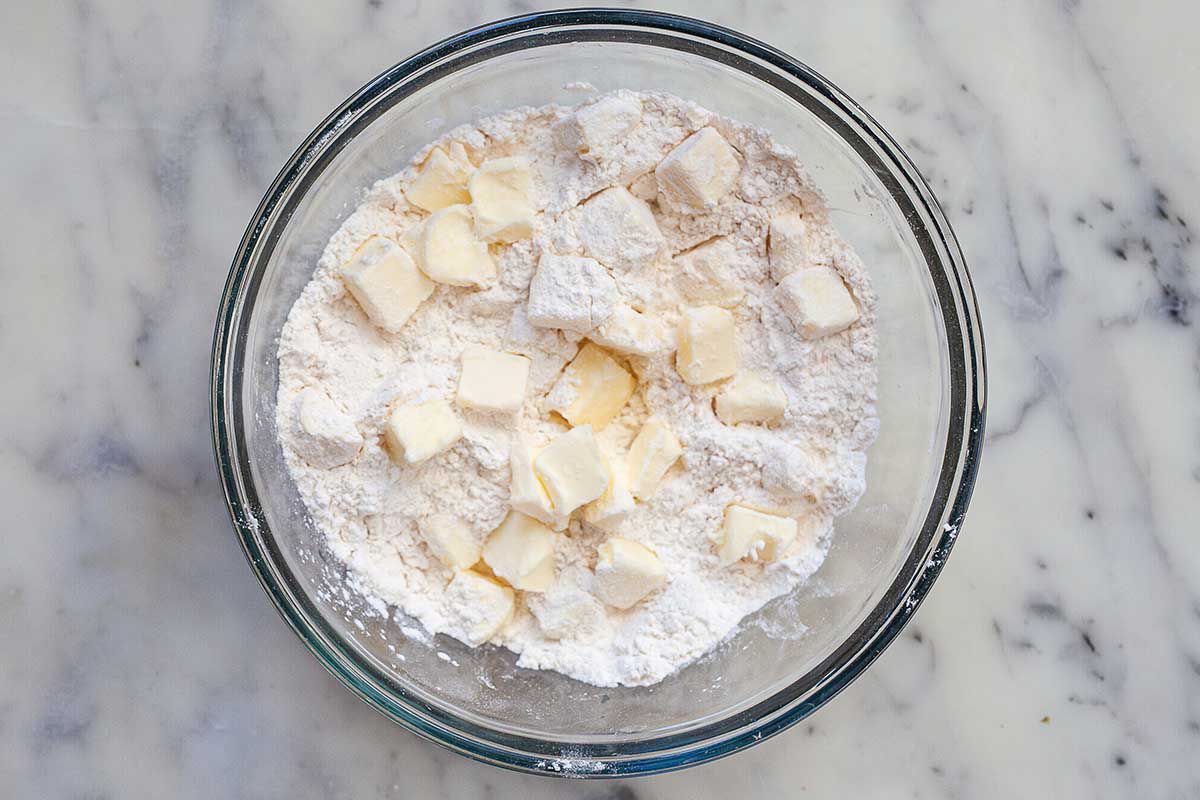Cubes of butter are in a flour mixture inside a glass bowl on a marble countertop.